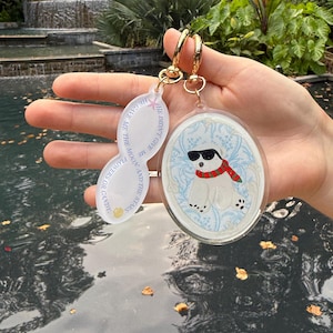 May include: Two keychains with gold-colored clasps. One keychain features an oval-shaped pendant with a polar bear wearing sunglasses and a red scarf. The other keychain has an oval pendant with the text: "HE GAVE ME THE MOON AND THE STARS, FLOWERS OR CANDY."