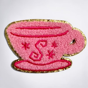May include: A pink tea cup patch with a textured, fuzzy surface and a gold glitter border. The cup features a dark pink embroidered letter S in the center, surrounded by small star-shaped accents, and a dark pink rim and base outline.