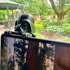 Puede incluir: Una figura de pl&aacute;stico negro de Darth Vader dise&ntilde;ada para engancharse en el borde superior de una pantalla. La figura presenta el ic&oacute;nico casco, la m&aacute;scara y manos enguantadas que descansan sobre el marco, con un acabado mate y l&iacute;neas de impresi&oacute;n 3D visibles.