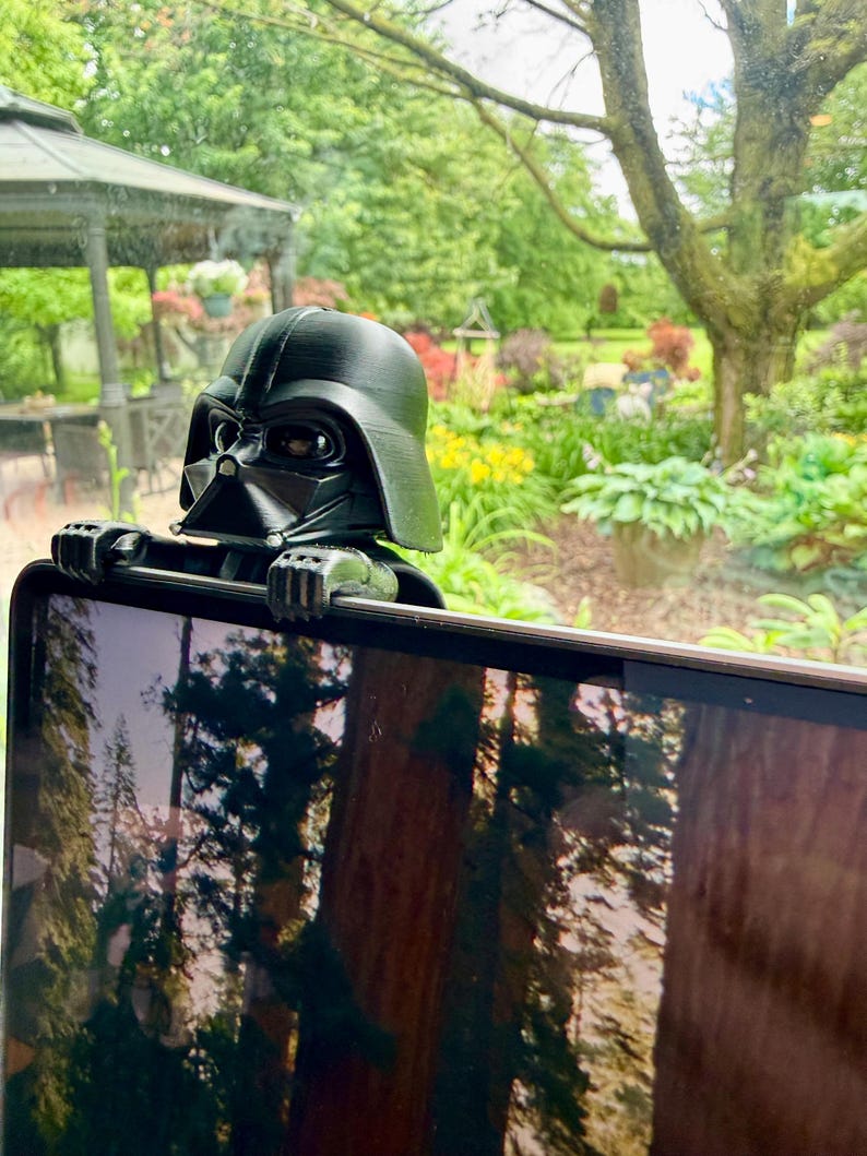 Puede incluir: Una figura de pl&aacute;stico negro de Darth Vader dise&ntilde;ada para engancharse en el borde superior de una pantalla. La figura presenta el ic&oacute;nico casco, la m&aacute;scara y manos enguantadas que descansan sobre el marco, con un acabado mate y l&iacute;neas de impresi&oacute;n 3D visibles.