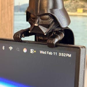 Puede incluir: Una figura de pl&aacute;stico negro de un personaje con casco y capa, posada en el borde superior de un monitor de computadora. La pantalla muestra una barra de estado con iconos y el texto Wed Feb 11 3:52 PM. La figura tiene un acabado liso.