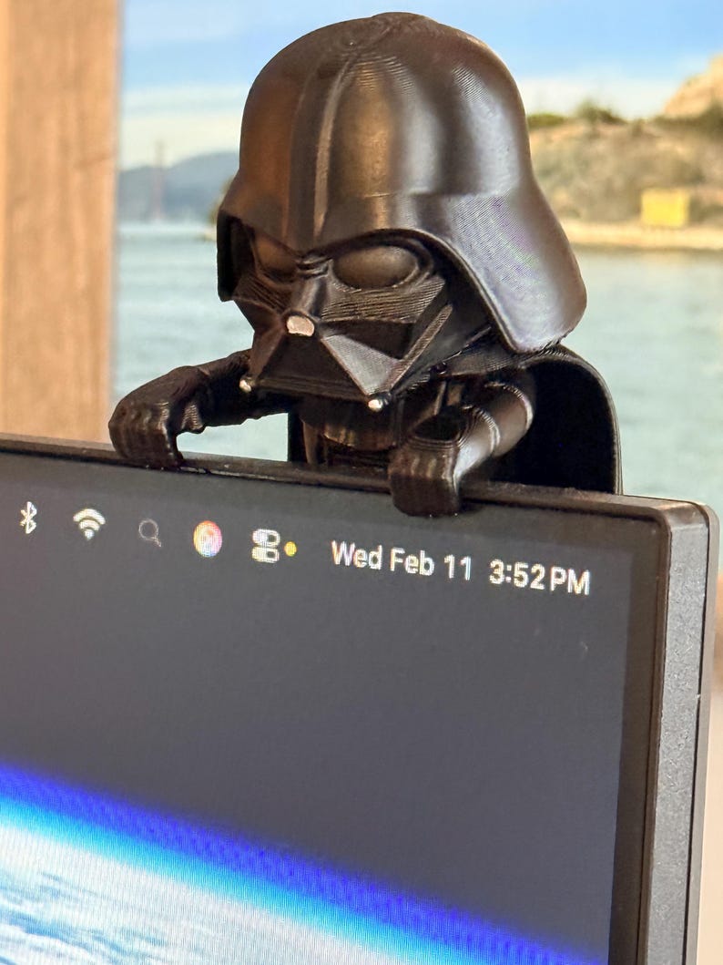 Puede incluir: Una figura de pl&aacute;stico negro de un personaje con casco y capa, posada en el borde superior de un monitor de computadora. La pantalla muestra una barra de estado con iconos y el texto Wed Feb 11 3:52 PM. La figura tiene un acabado liso.