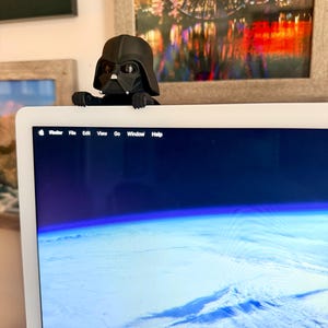 Puede incluir: Una figura negra de Darth Vader posada en el borde superior de un monitor de computadora. El personaje tiene un casco negro mate, una m&aacute;scara facial triangular con detalles plateados y manos peque&ntilde;as que sujetan el marco.