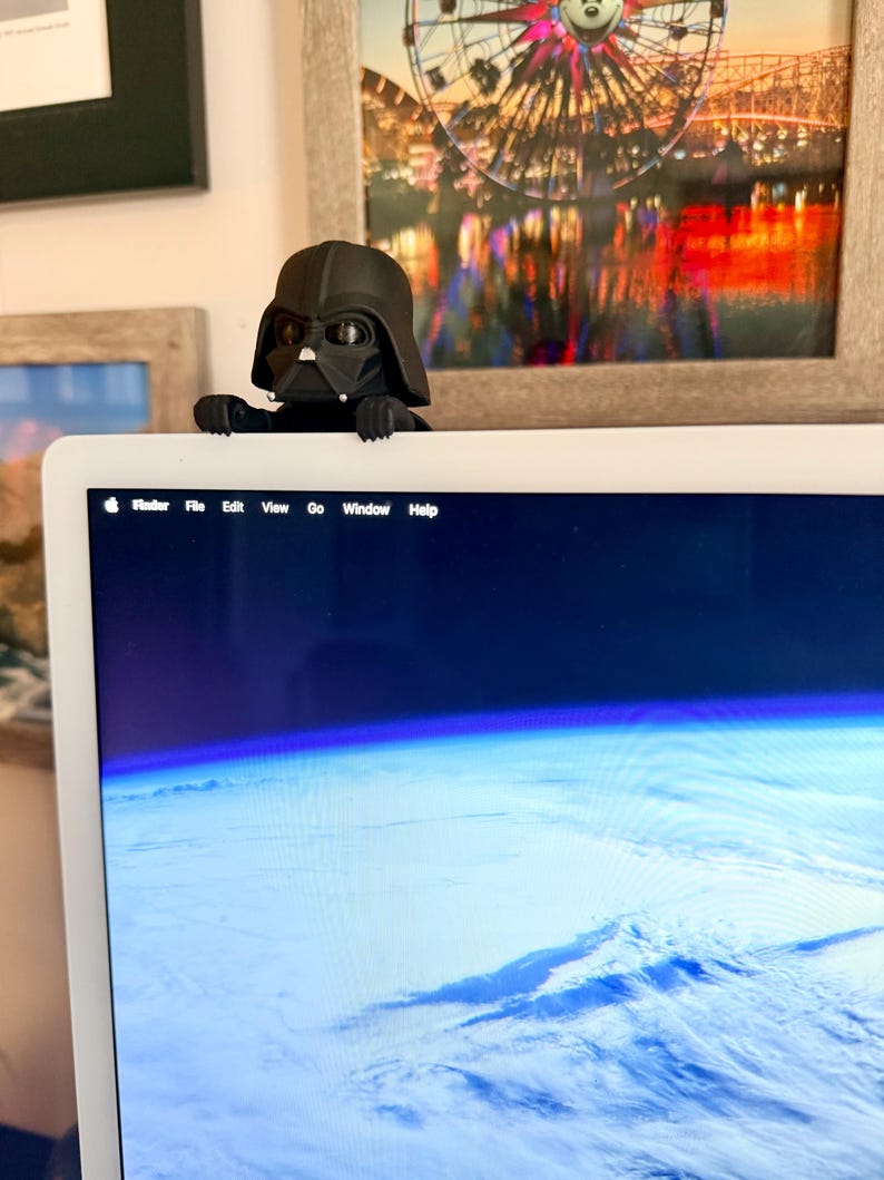 Puede incluir: Una figura negra de Darth Vader posada en el borde superior de un monitor de computadora. El personaje tiene un casco negro mate, una m&aacute;scara facial triangular con detalles plateados y manos peque&ntilde;as que sujetan el marco.