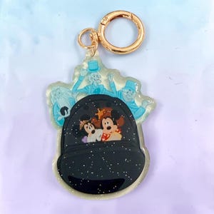 May include: A clear acrylic keychain featuring a gold-toned metal clasp. The design depicts a black, glittery vehicle carrying two cartoon mice wearing crowns, with three pale blue, translucent ghost figures positioned behind the vehicle.