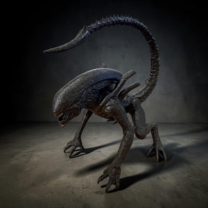 May include: A detailed sculpture of an Alien creature, featuring a dark, textured surface. The creature is posed on all fours with a long, curved tail and a distinctive head shape. The sculpture is set against a dark, textured background.