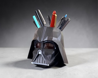 May include: A black, geometric, low-poly style desk organizer shaped like a helmet. The hollow top holds several pens and pencils, including a prominent red pen, a light blue pen, and various black writing instruments, all stored upright.