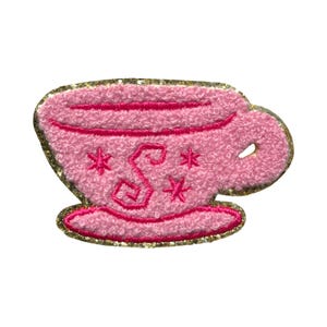 May include: Pink embroidered patch in the shape of a teacup. The teacup has a gold glitter border and features the letter "S" and starburst designs. The teacup sits on a matching pink saucer.