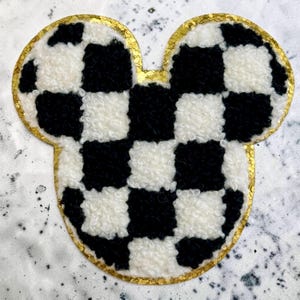 Peut inclure: Un écusson en forme de souris avec un motif à damier noir et blanc en matière texturée et duveteuse. La forme entière est soulignée par une épaisse bordure dorée scintillante qui ajoute un contraste métallique à la surface en tissu doux.