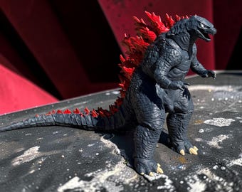 May include: A dark gray Godzilla action figure with textured, reptilian skin and prominent, jagged red dorsal fins running along its back and tail. The creature stands on two legs with sharp, light-colored claws on its feet and hands.