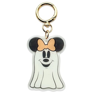 May include: A clear, glittery Minnie Mouse ghost keychain with a gold-colored clasp. The ghost has black ears, a peach bow, and a smiling face. The keychain is a fun, festive accessory.
