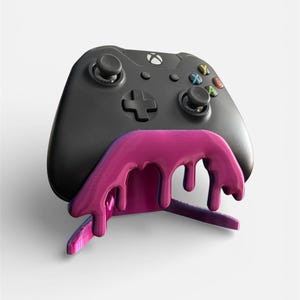 May include: A black Xbox controller rests on a vibrant pink and purple stand. The stand has a unique design with a dripping paint effect, adding a creative touch to the gaming accessory. The controller features colored buttons and a directional pad.
