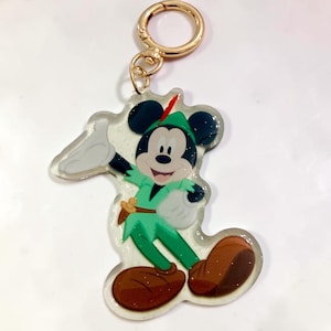 May include: A clear acrylic keychain featuring a graphic of Mickey Mouse dressed as Peter Pan in a green tunic, brown shoes, and a cap with a red feather. The character is depicted with white gloves, and the keychain includes a gold-toned metal ring.
