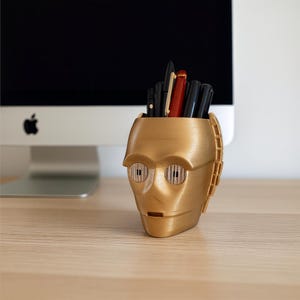 May include: A gold-colored pen holder shaped like a robot's head, with cylindrical eye details and a rectangular mouth. The pen holder is filled with various pens and pencils. The background includes a computer monitor and a wooden desk.