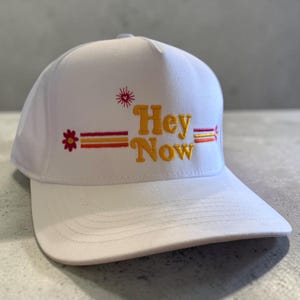 May include: White baseball cap with the embroidered text "Hey Now" in orange and yellow, with a flower and star design. The cap has a flat brim and a structured crown.