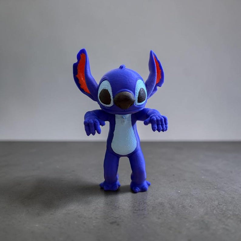 May include: A blue figurine of the character Stitch from the Disney movie Lilo & Stitch. The figurine has large ears with red accents, a brown nose, and large black eyes. The figure is standing with its arms outstretched.