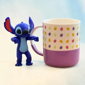 May include: A blue Stitch figurine stands next to a ceramic mug. The mug has a white upper section with colorful polka dots and a purple lower section. The handle and the base of the mug are also purple.
