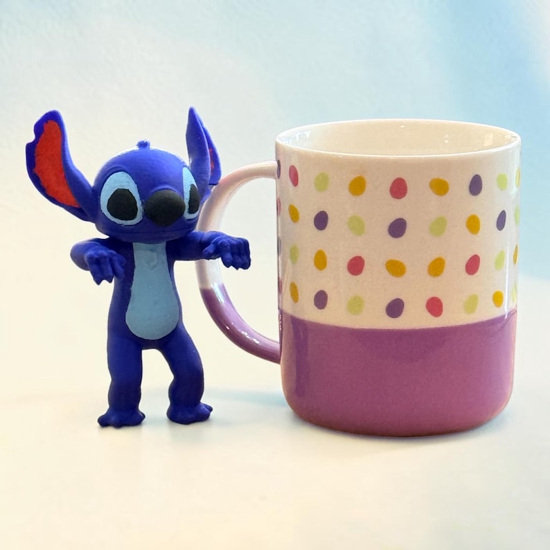May include: A blue Stitch figurine stands next to a ceramic mug. The mug has a white upper section with colorful polka dots and a purple lower section. The handle and the base of the mug are also purple.