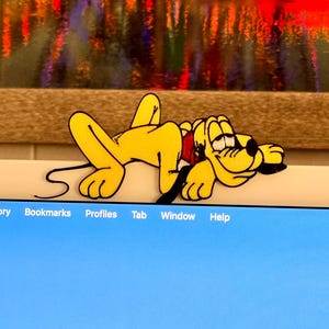 May include: A yellow cartoon dog figure, with black outlines and a red collar, is lying down on a light blue surface. The dog is in a relaxed pose, with its eyes closed. The background features a blurred, colorful image.