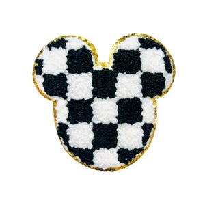 May include: A Mickey Mouse-shaped patch featuring a black and white chequered pattern. The patch has a gold glitter border and a fluffy, textured appearance. The design is reminiscent of a racing flag.