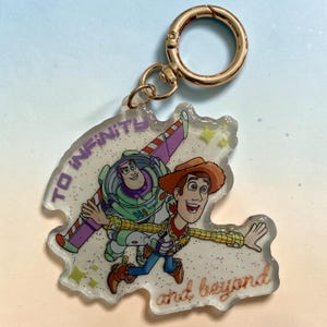 May include: A clear acrylic keychain featuring a printed illustration of Buzz Lightyear and Woody from Toy Story. The design includes the purple text TO INFINITY and the brown cursive text and beyond. It has a gold-toned metal lobster clasp.