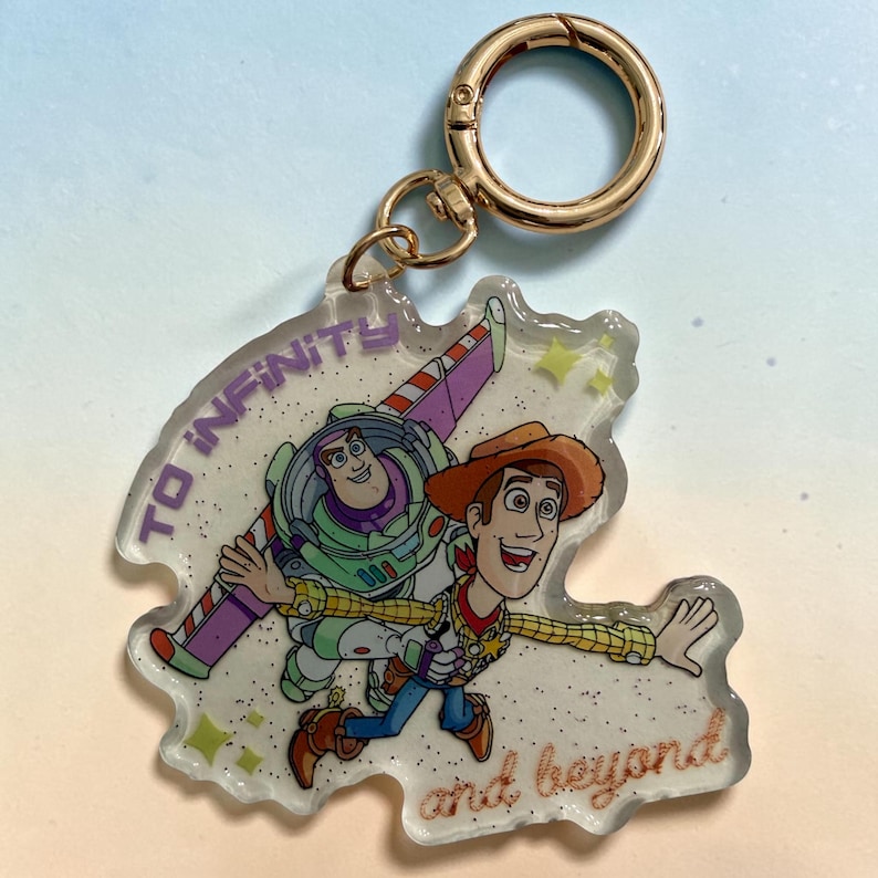 May include: A clear acrylic keychain featuring a printed illustration of Buzz Lightyear and Woody from Toy Story. The design includes the purple text TO INFINITY and the brown cursive text and beyond. It has a gold-toned metal lobster clasp.