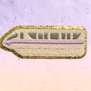 May include: A rectangular embroidered patch featuring a cream-colored monorail train with grey windows and a horizontal lavender stripe. The patch is outlined with a thick, shimmering gold glitter border, creating a distinct contrast against the textured fabric.