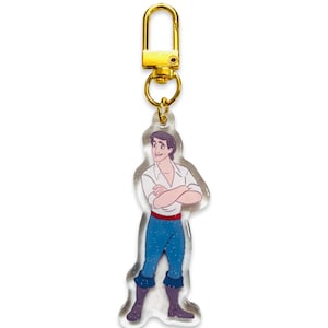 May include: A clear acrylic keyring featuring a cartoon illustration of a character in a white shirt, blue trousers, and brown boots. The keyring has a gold-coloured clasp.