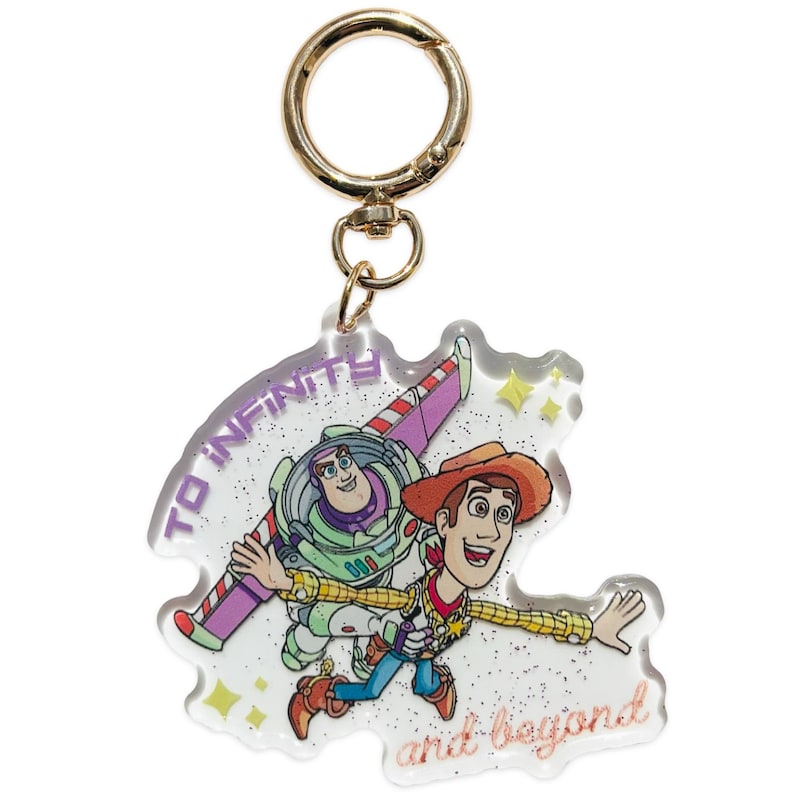 May include: A clear acrylic keychain featuring a printed illustration of Buzz Lightyear and Woody from Toy Story. The design includes the purple text TO INFINITY and the orange text and beyond, with gold glitter accents and a gold metal keyring.