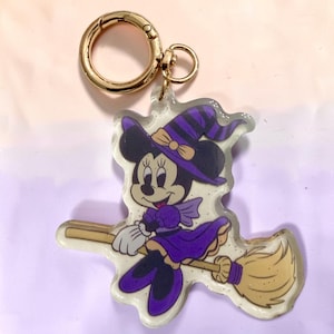 May include: A clear acrylic keychain featuring a cartoon mouse character dressed as a witch in a purple and black striped hat and purple dress, riding a tan broomstick. The design includes gold glitter accents and a gold-toned metal lobster clasp.