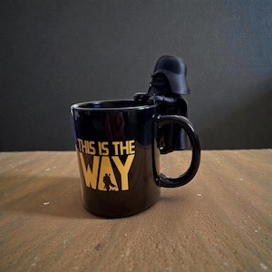 Puede incluir: Una taza de cer&aacute;mica negra con el texto dorado THIS IS THE WAY y una peque&ntilde;a silueta de un personaje. Una figura moldeada negra de Darth Vader est&aacute; unida al borde de la taza, colocada directamente sobre el asa, mirando hacia afuera.