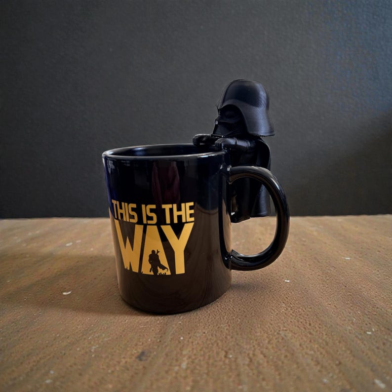 Puede incluir: Una taza de cer&aacute;mica negra con el texto dorado THIS IS THE WAY y una peque&ntilde;a silueta de un personaje. Una figura moldeada negra de Darth Vader est&aacute; unida al borde de la taza, colocada directamente sobre el asa, mirando hacia afuera.