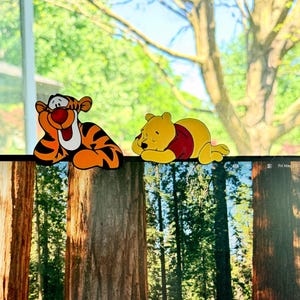 May include: Two cartoon character cutouts, Tigger and Winnie the Pooh, are displayed. Tigger is orange with black stripes, and Winnie the Pooh is yellow with a red shirt. The background shows a window with trees.