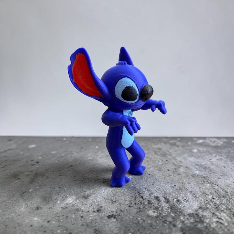 May include: A blue figurine of the character Stitch from the animated film Lilo & Stitch. The figurine has large ears with red interiors, black eyes, and a light blue belly. It is posed with its arms outstretched.