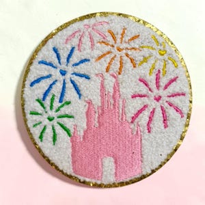 May include: A circular embroidered patch featuring a white textured background with a pink castle silhouette in the center. Surrounding the castle are colorful stitched fireworks in blue, green, orange, yellow, and pink. The edge has a gold border.