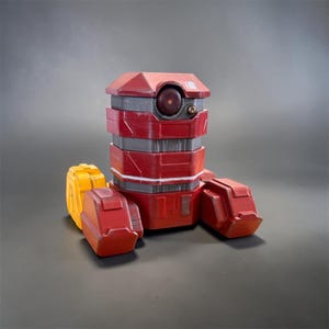 May include: A robotic figure featuring a cylindrical red and grey body with a single dark circular lens on the front. The base includes three blocky feet, two of which are red and one of which is yellow, with geometric paneling details.