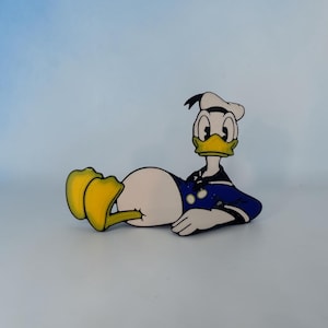 May include: A wooden cutout of Donald Duck in a relaxed pose. The cartoon character is depicted with a white body, yellow feet, and a blue sailor suit. His iconic sailor hat is white, and his beak is yellow.