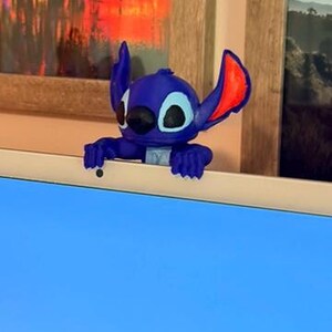 May include: A blue and red Stitch figurine, a character from the animated film Lilo & Stitch, is perched on a white surface. The figurine has large ears, black eyes, and a wide smile. The background includes a window and a framed picture.