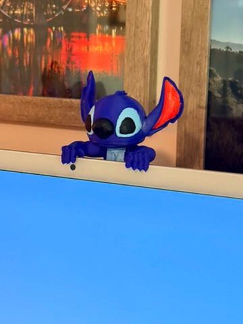 May include: A blue and red Stitch figurine, a character from the animated film Lilo & Stitch, is perched on a white surface. The figurine has large ears, black eyes, and a wide smile. The background includes a window and a framed picture.