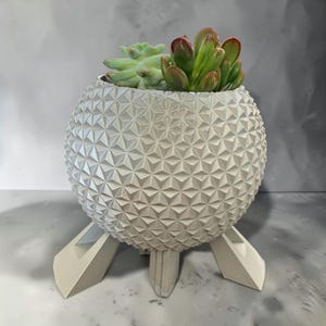 May include: A spherical off-white planter with a geometric triangular textured surface, supported by three angled legs. The planter contains two small succulent plants, one pale green and one with reddish-tipped green leaves.