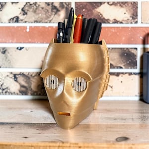 May include: A metallic gold-colored desk organizer shaped like a robot head with circular, ridged eye details and a small rectangular mouth opening. The hollow top section contains a collection of various black and red pens and pencils.