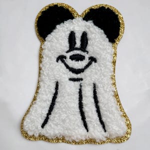 May include: A white, textured, fuzzy fabric patch shaped like a ghost with black embroidered ears, eyes, nose, and mouth. The patch features a gold glitter border that outlines the entire shape, creating a distinct contrast against the white.