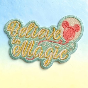 May include: An embroidered patch with a light green background featuring the gold cursive text Believe in Magic. A pink balloon shaped like a mouse head is attached to the text by a thin pink string, and small white star accents are scattered throughout.