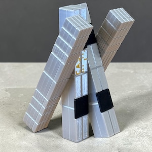 May include: A 3D-printed sculpture of a figure in a uniform. The figure is primarily light gray with black accents and gold detailing. The design features a structured, blocky aesthetic.
