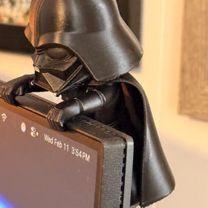 Puede incluir: Una figura negra impresa en 3D de Darth Vader posada en el borde superior de un monitor de computadora. El personaje se muestra con su caracter&iacute;stico casco, m&aacute;scara y capa fluida, con las manos apoyadas en el marco de la pantalla.