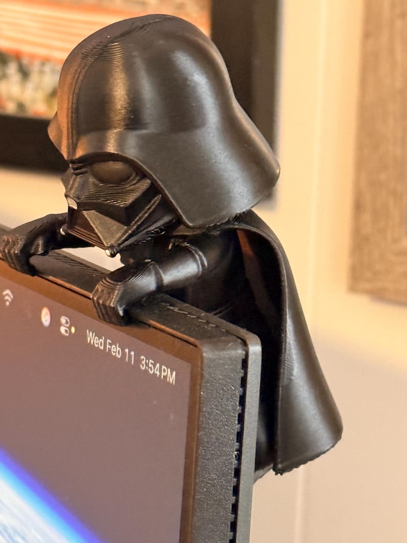 Puede incluir: Una figura negra impresa en 3D de Darth Vader posada en el borde superior de un monitor de computadora. El personaje se muestra con su caracter&iacute;stico casco, m&aacute;scara y capa fluida, con las manos apoyadas en el marco de la pantalla.