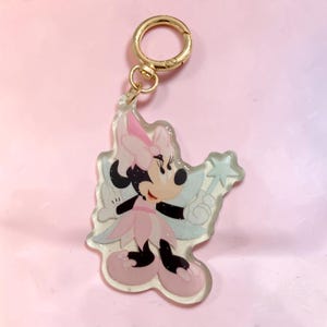 May include: A clear acrylic keychain featuring a cartoon mouse character dressed as a fairy in a pink dress and hat with light blue wings. The character holds a star-tipped wand. The keychain includes a gold-toned metal lobster clasp ring.