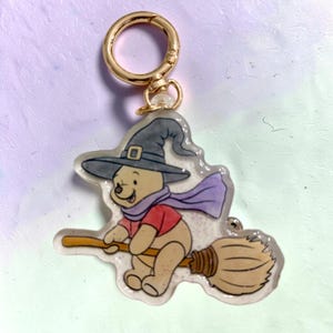 May include: A clear acrylic keychain featuring a cartoon bear wearing a dark gray witch hat and a purple scarf, riding a brown broomstick. The bear has a red shirt and light brown fur. A gold-toned metal lobster clasp is attached to the top.