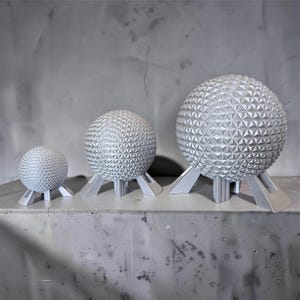 May include: Three silver-colored spherical sculptures of varying sizes, each featuring a geometric geodesic dome pattern composed of small triangles. Each sphere is supported by a set of three angled, sturdy legs that elevate the structure.