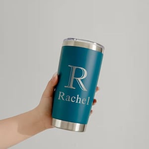 May include: Teal insulated tumbler with a silver lid and base. The tumbler features a large silver letter "R" and the name "Rachel" printed below. The tumbler is cylindrical and appears to be made of stainless steel.